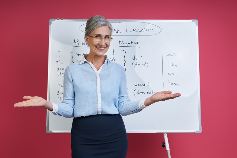 Cheerful senior woman teaching English language near the whiteboard against pink background